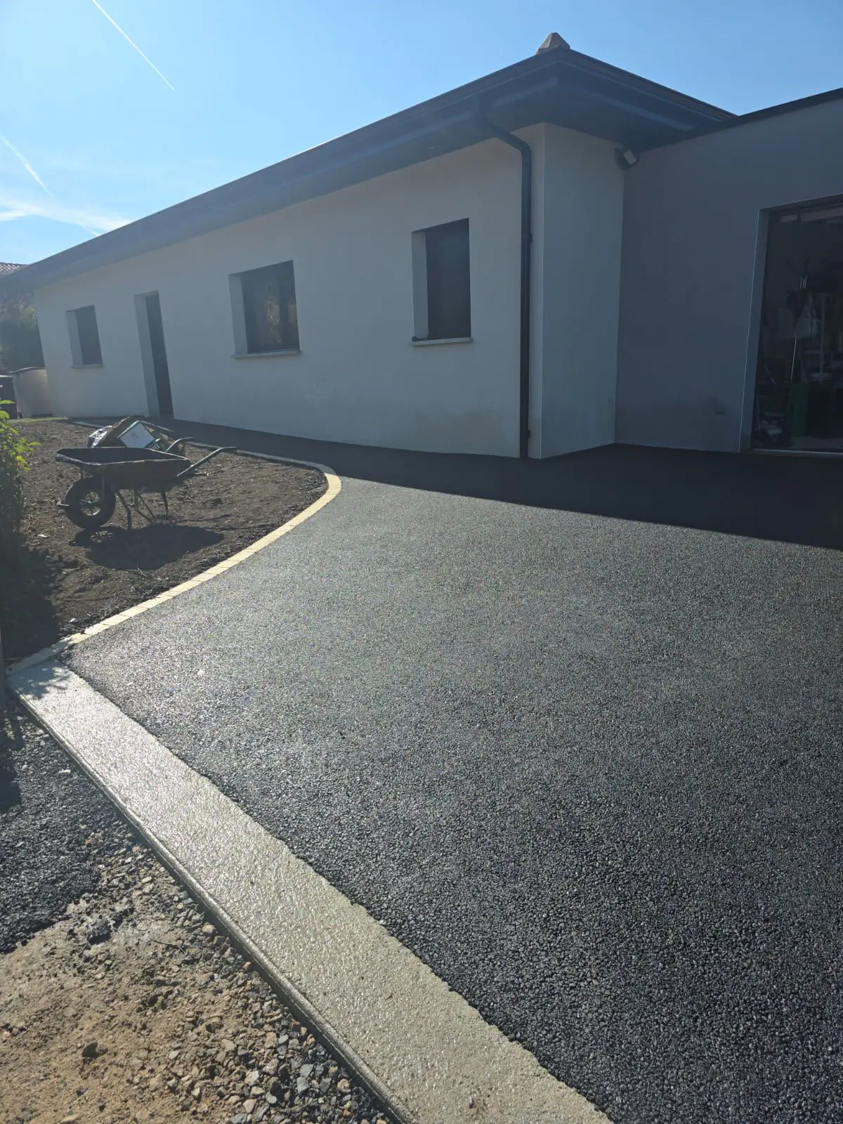 Cour en enrobé neuf soulignée d'une bordure béton courbe devant une maison plain-pied blanche
