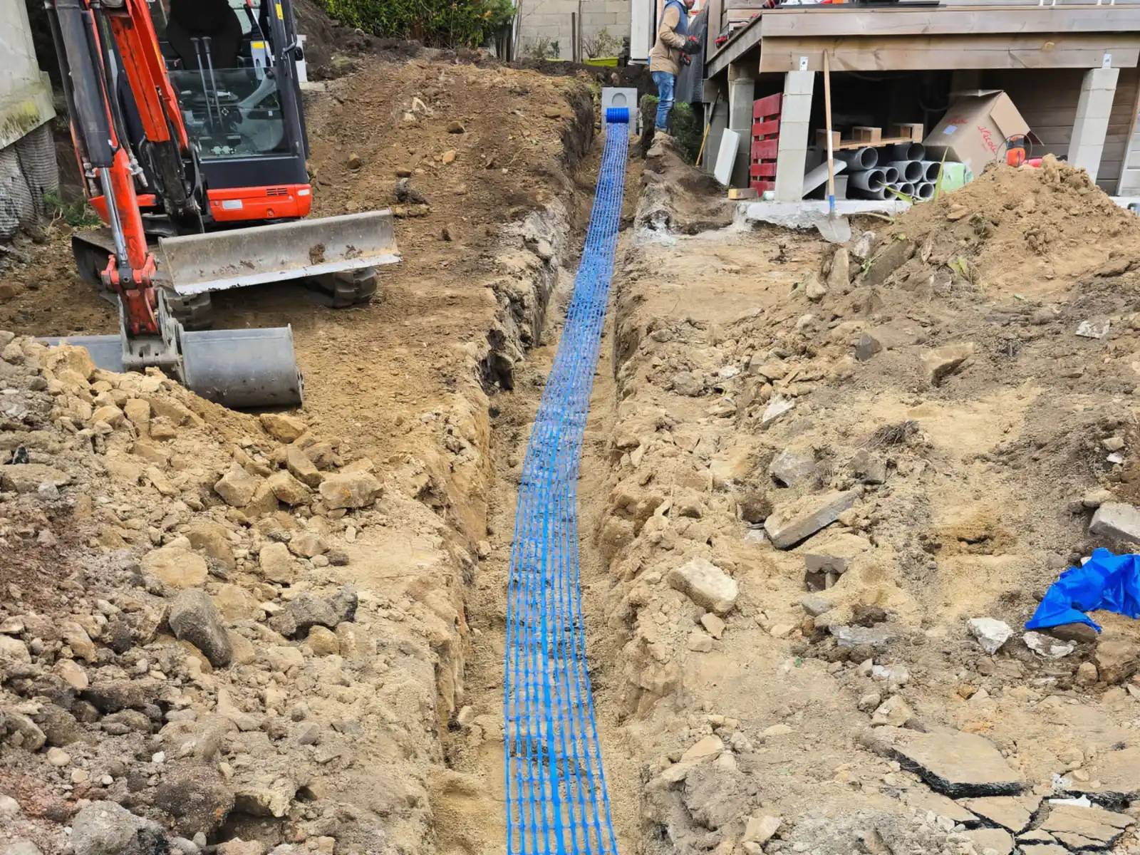 Tranchée de réseaux avec grillage avertisseur bleu, mini-pelle en action en arrière-plan